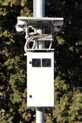 Four small white ball shaped rotatable waterproof security cameras CCTV powered by solar panel on top mounted on strong metal utility pole above processing unit cooled by two black fans with dense tre