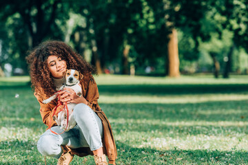 Curly woman in raincoat hugging jack russell terrier on lawn in park