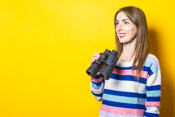 Young woman with a smile holds binoculars in her hands on a yellow background. Banner