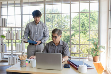 A middle-aged Asian man teaching his son a job standing next to a desk in an office.