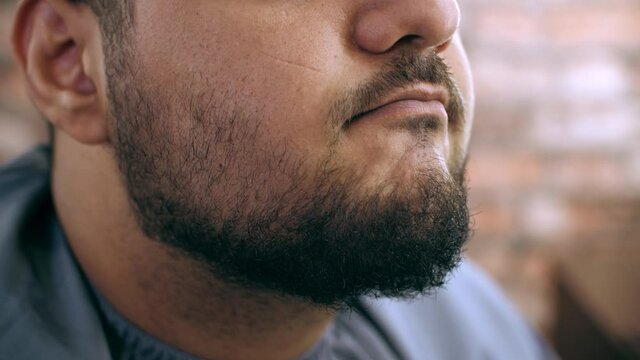 Close-up view of man touching shaved beard in barber shop. Cropped shot of handsome man in mantle checking facial hair after grooming in barbershop
