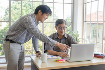 A middle-aged Asian man teaching his son a job standing next to a desk in an office.
