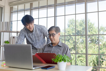 A middle-aged Asian man teaching his son a job standing next to a desk in an office.