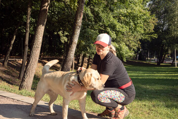 A young pregnant woman in sportswear and a baseball cap plays with a labrador dog in a city park. Warm sunny day, active lifestyle.