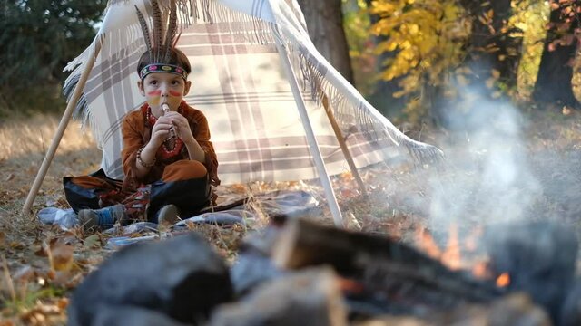 A little boy dressed in a Native American costume, sat down by the wigwam and played the flute. Playing with Indians near the fire with feathers on his head.