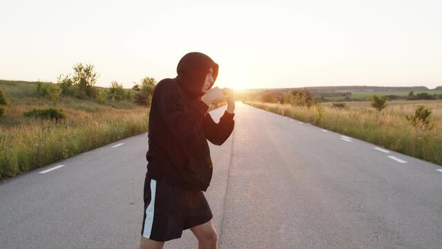 Kickboxer with hood on head training kicks on country road singly during sunset