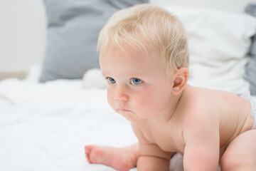 Ten month baby boy sitting on bed. Little naked kid with blue eyes in nappy at home. Baby care concept, banner copy space