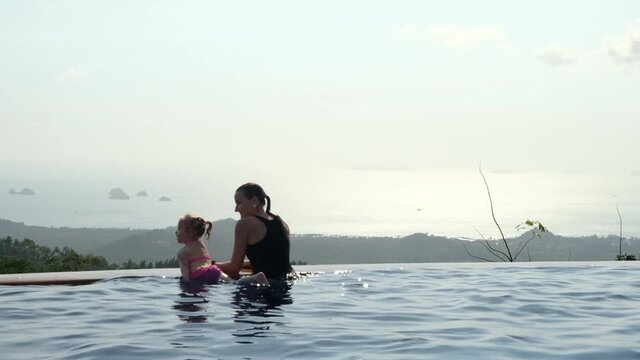 Young mother with daughter relaxing in infinity swimming pool looking at view