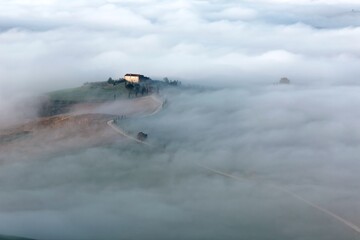 Aerial view of an isolated farmhouse on a foggy morning & a hilltop village in distant background ~ Amazing landscape of Tuscany countryside with rolling hills veiled in morning fog in Pienza, Italy