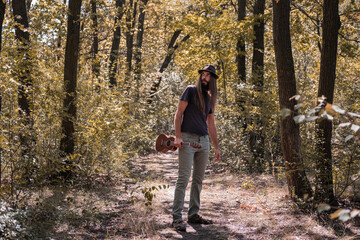 Fototapeta premium Bearded male with longhait and hat posing with ukulele in the forest 