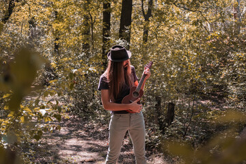 Bearded male with longhait and hat posing with ukulele in the forest 