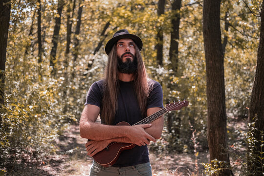 Bearded male with longhait and hat posing with ukulele in the forest 