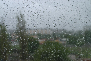 Wet glass with raindrops in cloudy weather on a blurry background of urban landscape, green trees and buildings