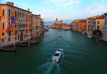 Fototapeta premium Romantic scenery of Venice at sunset, with boats, ferries & gondolas cruising on Grand Canal & the majestic landmark building Basilica di Santa Maria della Salute bathed in warm sunlight in background