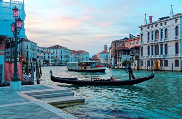 Scenery of romantic Venice at sunset, with view of a gondolier steering his ferry gondola by a pier, a vaporetto waterbus approaching from behind and beautiful old buildings by the Grand Canal © AaronPlayStation