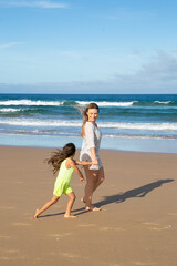 Fototapeta premium Happy mom holding little girls hand and walking on golden sand to sea, enjoying leisure time with daughter on beach. Full length. Family outdoor activities concept