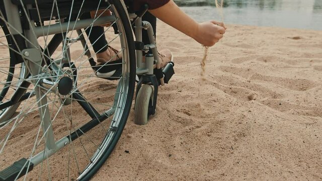close up, hand of disabled person in the wheelchair playing with the sand on the river coast. Vacation and travel with disability. High quality 4k footage