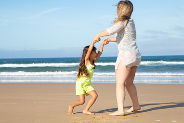 Cheerful adorable little girl and her mom having fun on beach, holding hands, dancing or spinning on sand. Full length. Family outdoor activities concept