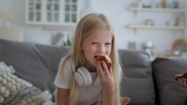 Portrait of young blonde beautiful girl with white headphones quenching her hunger holding delicious fresh cupcake cake and eating it while looking at camera operator sitting on sofa in living room.