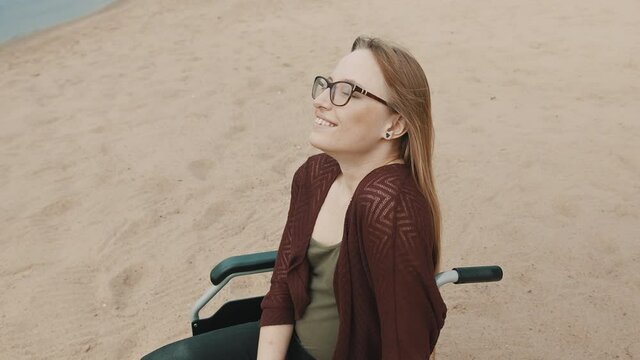 Young disabled woman on the sandy beach sitting in the wheelchair enjoying the sunny autumn day. High quality 4k footage