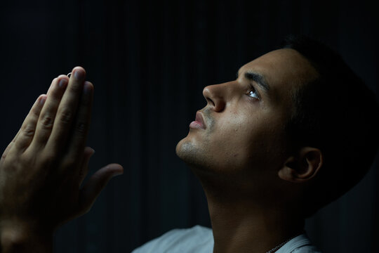 brunette boy in white t-shirt on black background praying. teenager looking at god with serious gaze and low light. man in silver necklace with serious expression while begging