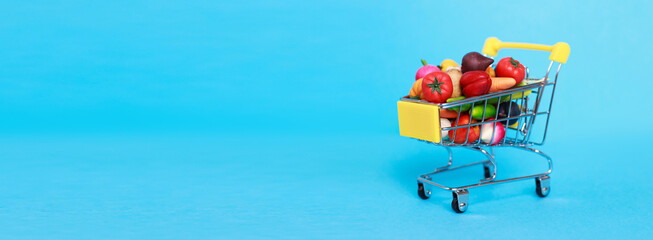 Fototapeta premium Metal shopping cart with fruits and vegetables on a blue background. Toy miniature shopping trolley