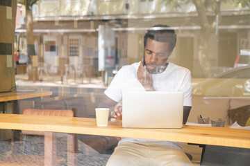 Serious African American man typing on laptop and speaking on cellphone in co-working space. View from street through window glass. Communication concept