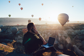 Tourist with laptop speaking on smartphone near hot air balloons