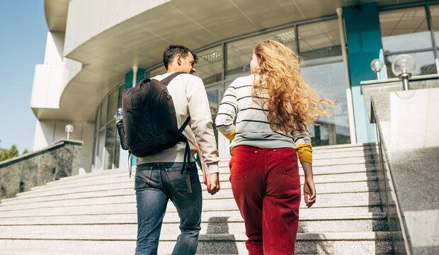 Rear view image of two young students talking to each other during climbing the stairs at the university outside. Two colleagues, a young woman and a man going to the college campus together.