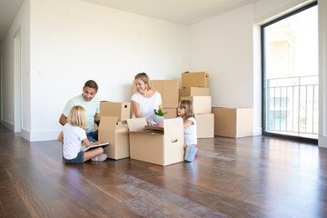 Happy parents and two kids moving into new empty apartment, sitting on floor near open boxes. Wide shot. Relocation or moving concept