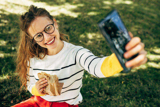 Smiling student female taking selfies on the mobile phone during lunch with healthy food sitting on the green grass at college campus. A gorgeous young woman has online conversations on the smartphone