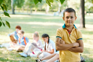 Portrait of smiling confident boy in yellow tshirt standing with crossed arms in summer park, student doing task in background