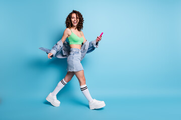 Fototapeta na wymiar Full length photo of positive cheerful girl go walk copyspace use smartphone enjoy social network chatting wear green crop top style stylish outfit isolated blue color background
