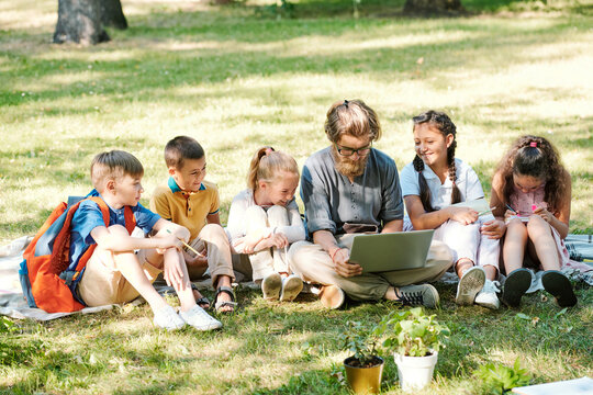 Hipster bearded teacher sitting on grass and using laptop while preparing project with schoolchildren in park