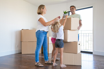 Fototapeta na wymiar Happy young family with moving boxes in their new house. Mom, dad and children standing near cardboard boxes, smiling. Little girl holding plant in pot. Mortgage, relocation and moving day concept