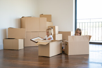 Focused little kids unpacking things in new apartment, sitting on floor and taking objects from open cartoon boxes. Relocation or moving concept