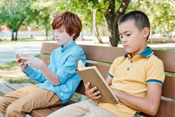 Redhead boys playing online game on smartphone while his friend sitting near him reading book