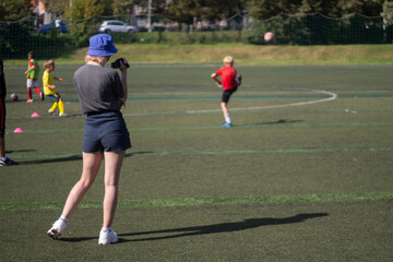 The girl takes pictures of sports. The girl is filming a football match. Girl in shorts on the playground.