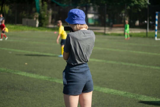 The girl takes pictures of sports. The girl is filming a football match. Girl in shorts on the playground.