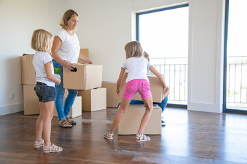 Happy family with two kids holding cardboard boxes and running into new home. Cute little girls helping parents with belongings in big apartment or house. Mortgage, relocation and moving day concept