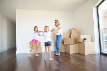 Fototapeta na wymiar Happy mother round dancing with two girls among unpacked boxes. Caucasian blonde mom having fan, smiling and playing with lovely daughters during removal. Family, relocation and moving day concept