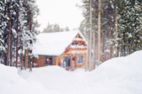 Defocused image of a cabin surrounded by deep snow and pine trees with falling snow and festive holiday lights