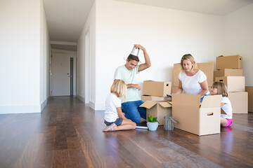 Cheerful parents and little daughters having fun while unpacking things in new apartment, sitting on floor and taking objects from open boxes. Relocation or moving concept