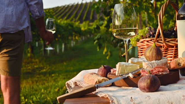 The man pours white wine into glasses. Beautiful people having romantic lunch with lots of tasty food and wine, sitting together on the picnic blanket at the vineyard on a sunny day. 4k footage