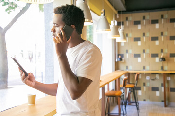 Serious African American guy talking on cell and looking at tablet screen. Young man using gadgets in co-working space or coffee shop. Digital communication concept