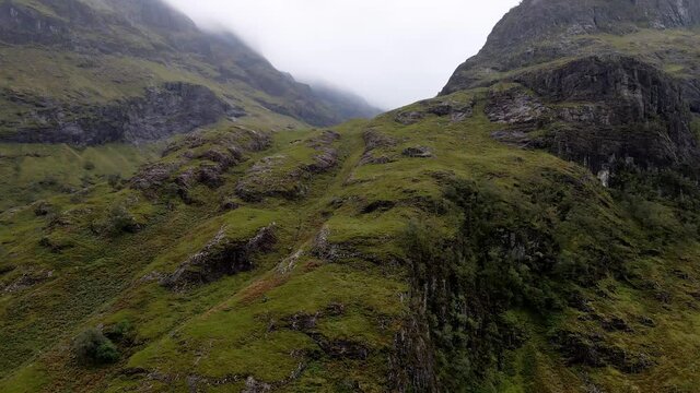 A drone shot revealing the sandy Glen Coe's canyon hidden in between rocky and cloudy Scottish highland's mountains
