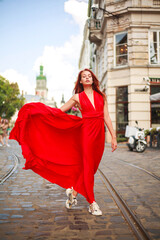 Fototapeta premium Attractive young woman in long red dress is walking along the square of tourist town