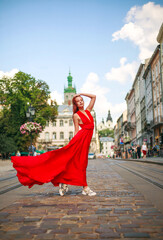 Naklejka premium Attractive young woman in long red dress is walking along the square of tourist town