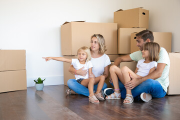 Fototapeta na wymiar Smiling little girls sitting on parents legs. Lovely daughter pointing away. Happy family sitting on floor together during relocation near carton boxes. Mortgage, property and moving day concept