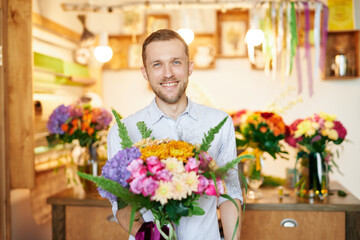Young beautiful smiling man with bouquet in flower shop. Good looking florist man in shirt holding bouquet with wide smile. Business concept.
